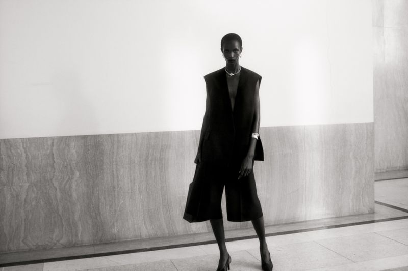 Elegant black and white fashion shot of a slender woman in a dark sleeveless jacket and culottes posing against a plain wall.