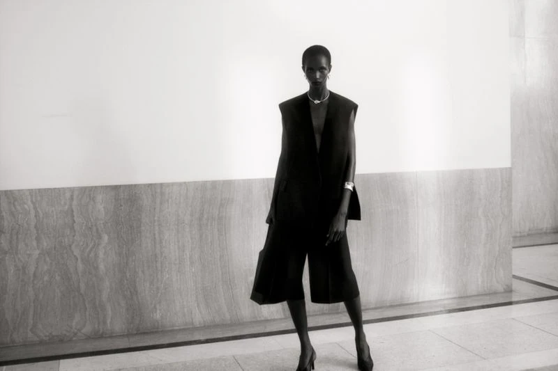 Elegant black and white fashion shot of a slender woman in a dark sleeveless jacket and culottes posing against a plain wall.