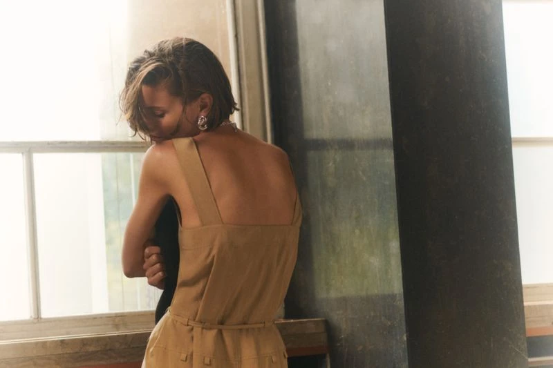 Back view of a short-haired woman in a sleeveless dress, with silver hoop earrings, posing in front of a window in a studio.