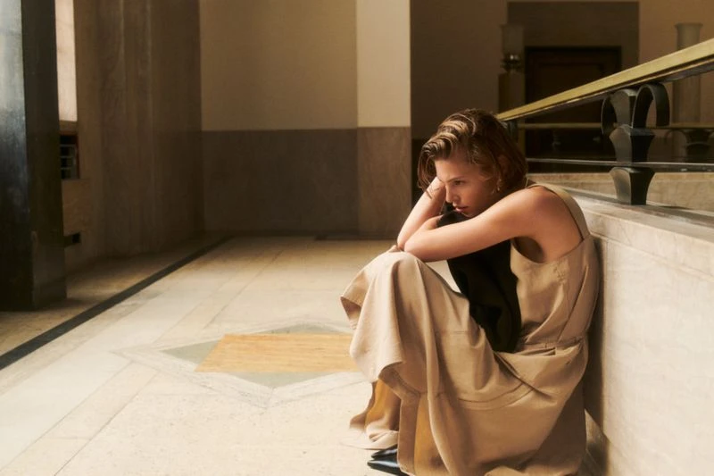 Adult woman in a tan dress sits on the marble floor indoors with her head resting in her hands near a wrought-iron railing.
