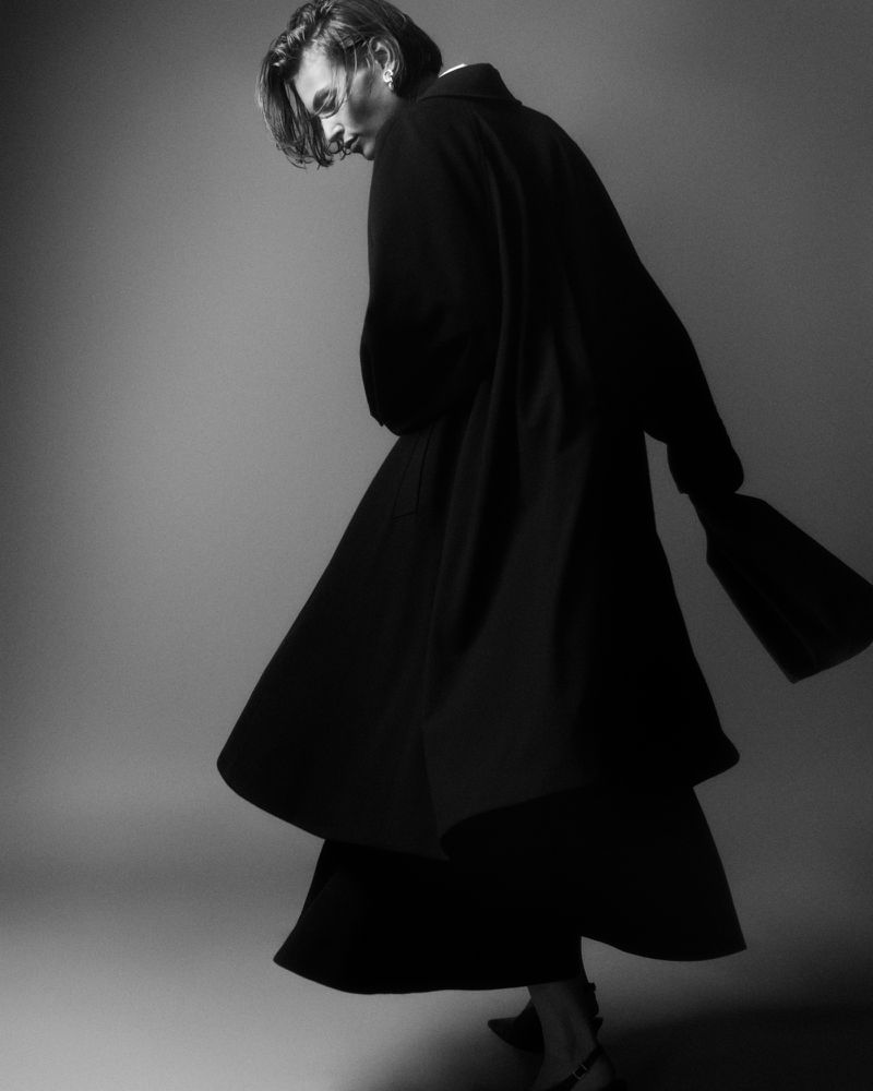 Model in a black coat holding a handbag against a seamless grey backdrop