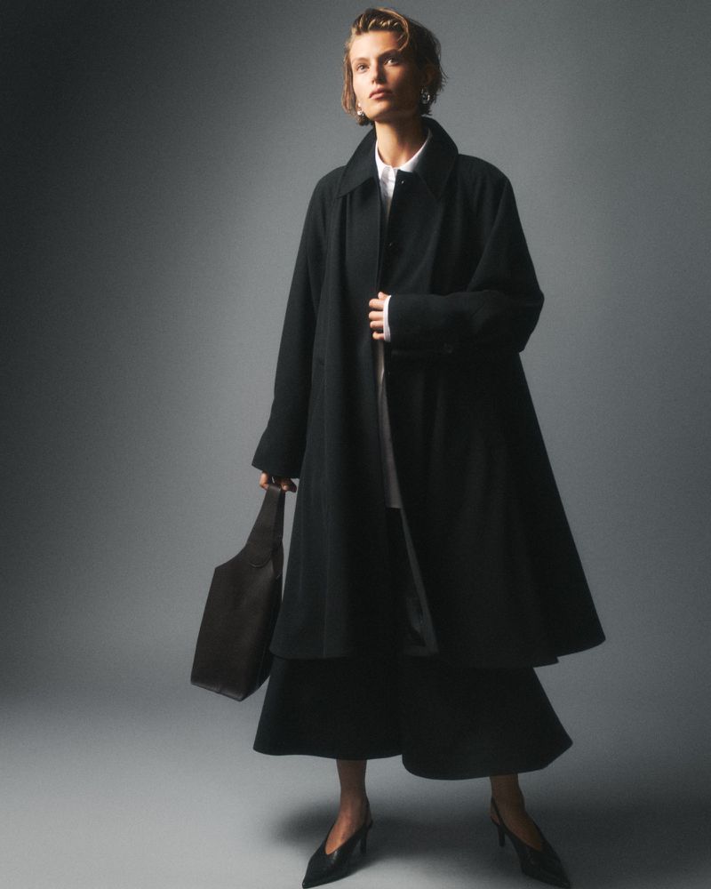 A woman in a dark coat and skirt stands in a studio with a black handbag and high heels. The image is in muted colors.