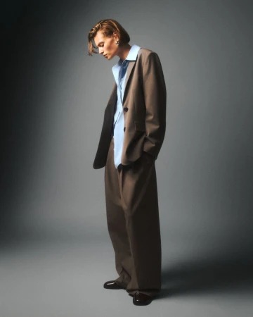 Full-length portrait of a person wearing a brown suit with an unbuttoned light blue shirt looking down against a grey backdrop.
