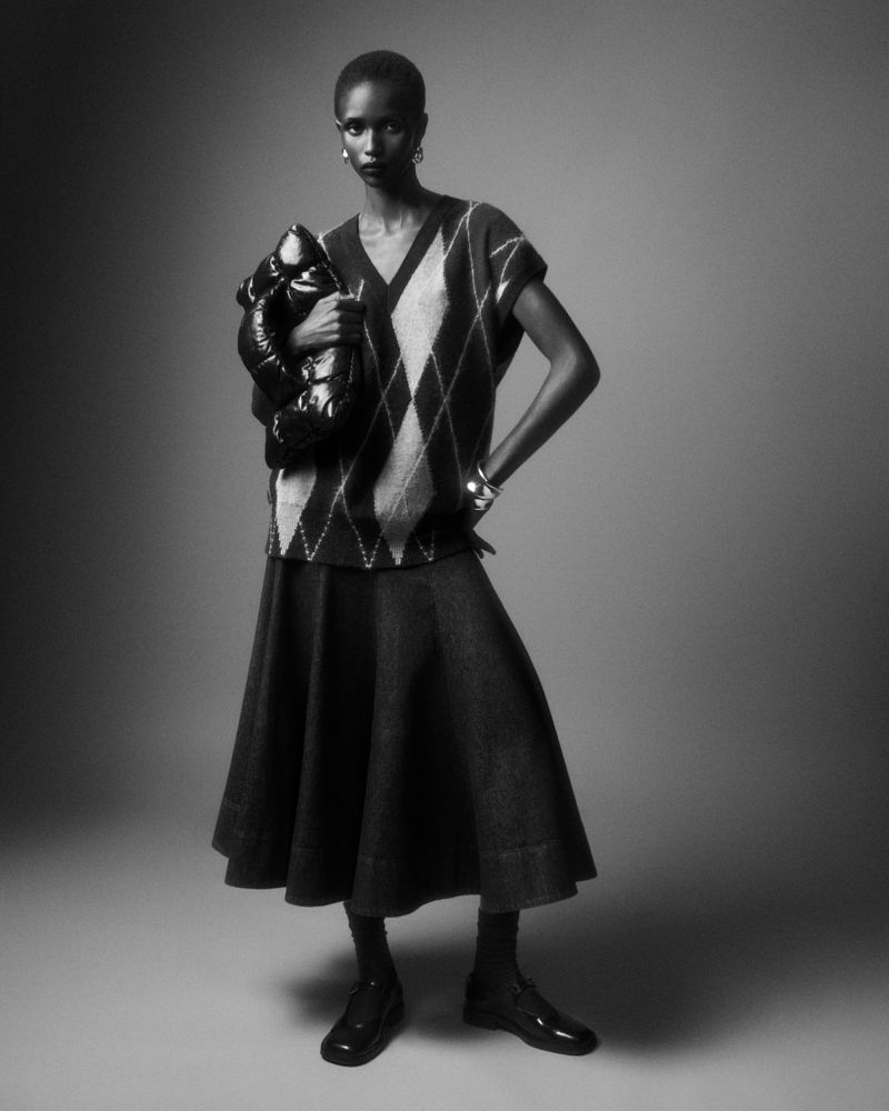 Black and white portrait of a Black woman in a patterned vest, skirt, and dress shoes holding a black puffer handbag.