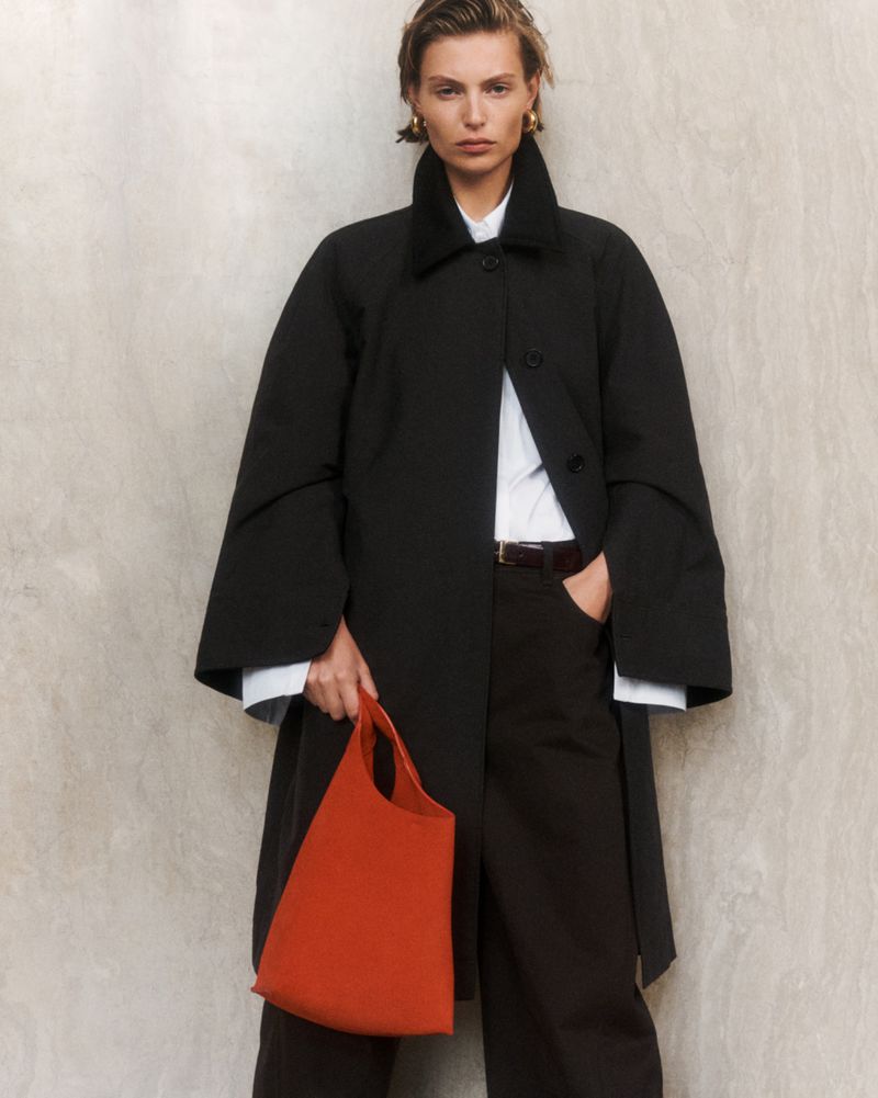 A female fashion model poses in a studio wearing a black coat, black pants, a white shirt, and holding an orange tote bag.