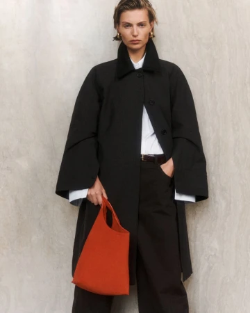 A female fashion model poses in a studio wearing a black coat, black pants, a white shirt, and holding an orange tote bag.