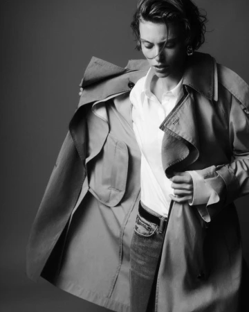 Model in a white shirt and trench coat posing against a seamless dark studio background.