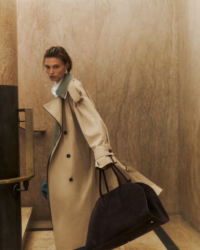 Model in a long beige trench coat holding a dark brown handbag inside a marble-walled interior with stairs.