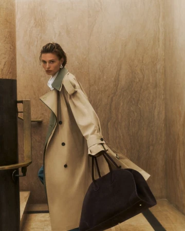 Model in a long beige trench coat holding a dark brown handbag inside a marble-walled interior with stairs.