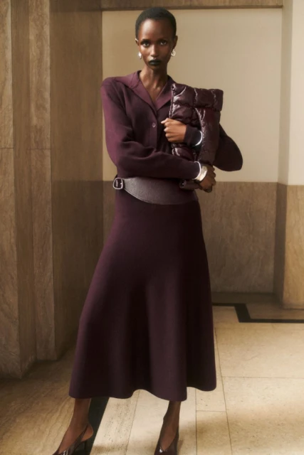 Elegant Black woman in a plum knit dress and heels holds a matching puffy bag, posing against a muted, neutral architectural interior.