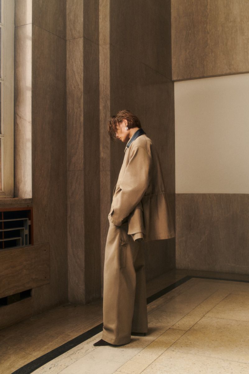An adult leaning in a modern corridor wearing an oversized tan suit, his head bowed, photographed in an architectural space.