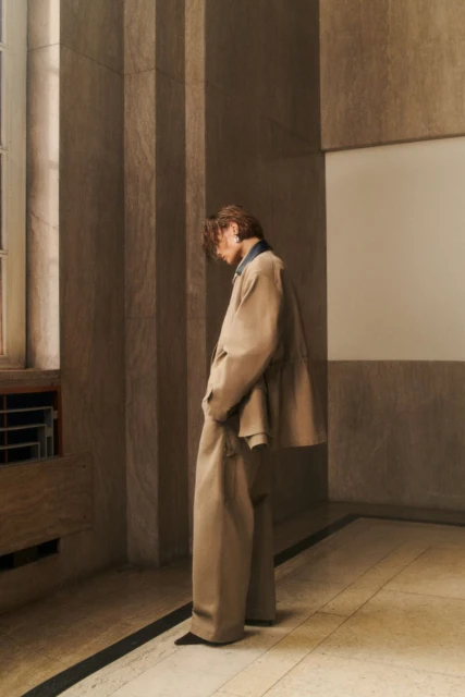 An adult leaning in a modern corridor wearing an oversized tan suit, his head bowed, photographed in an architectural space.