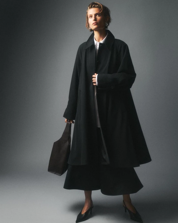 Model in a black coat wearing a white shirt and holding a dark handbag against a grey studio backdrop