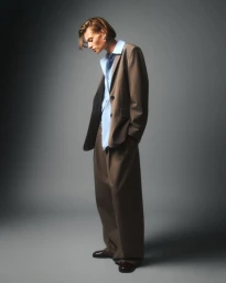 Model wearing an oversized brown suit jacket over a light blue buttoned shirt against grey seamless backdrop
