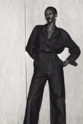 Monochrome portrait of a model wearing a dark denim button-down shirt and matching trousers against a pale textured background.