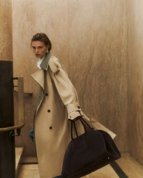 Model in a long beige trench coat holding a dark brown handbag inside a marble-walled interior with stairs.