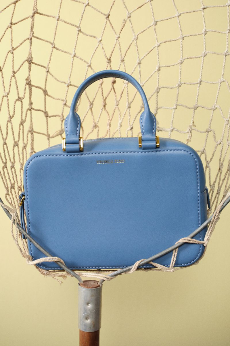 Studio shot of a light blue, rectangular handbag with rounded edges, featuring a top handle and gold hardware, against a yellow backdrop.
