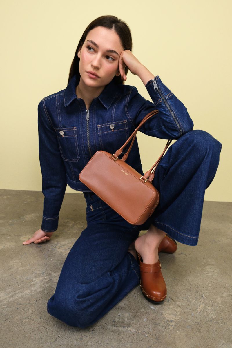 A woman in a denim outfit poses with a brown leather handbag and brown clogs against a yellow background in a studio setting.