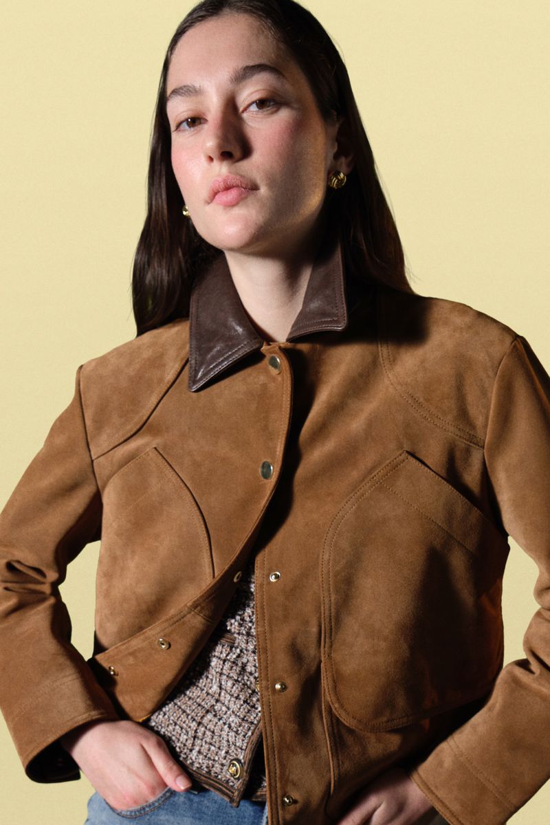 Close-up portrait of a young woman with brown hair wearing a brown suede jacket against a pastel yellow background.