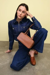 Model kneeling in dark denim jacket and matching pants holding a brown leather handbag against a pale yellow wall.