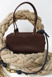 Dark brown leather handbag resting against a thick coiled rope prop on a white background