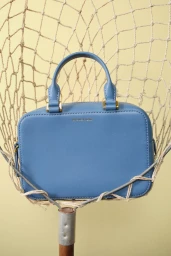 A light blue leather top-handle handbag resting inside a woven mesh net against a pale yellow background.