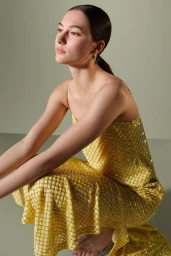Model crouching in a shimmering yellow spaghetti-strap dress and clear heels against a pale green-grey studio backdrop.