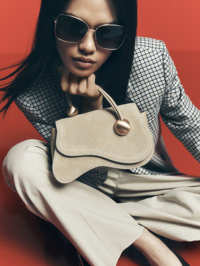 Stylish woman poses indoors with a unique tan handbag, wearing sunglasses, a checkered jacket, and neutral pants on a red backdrop.