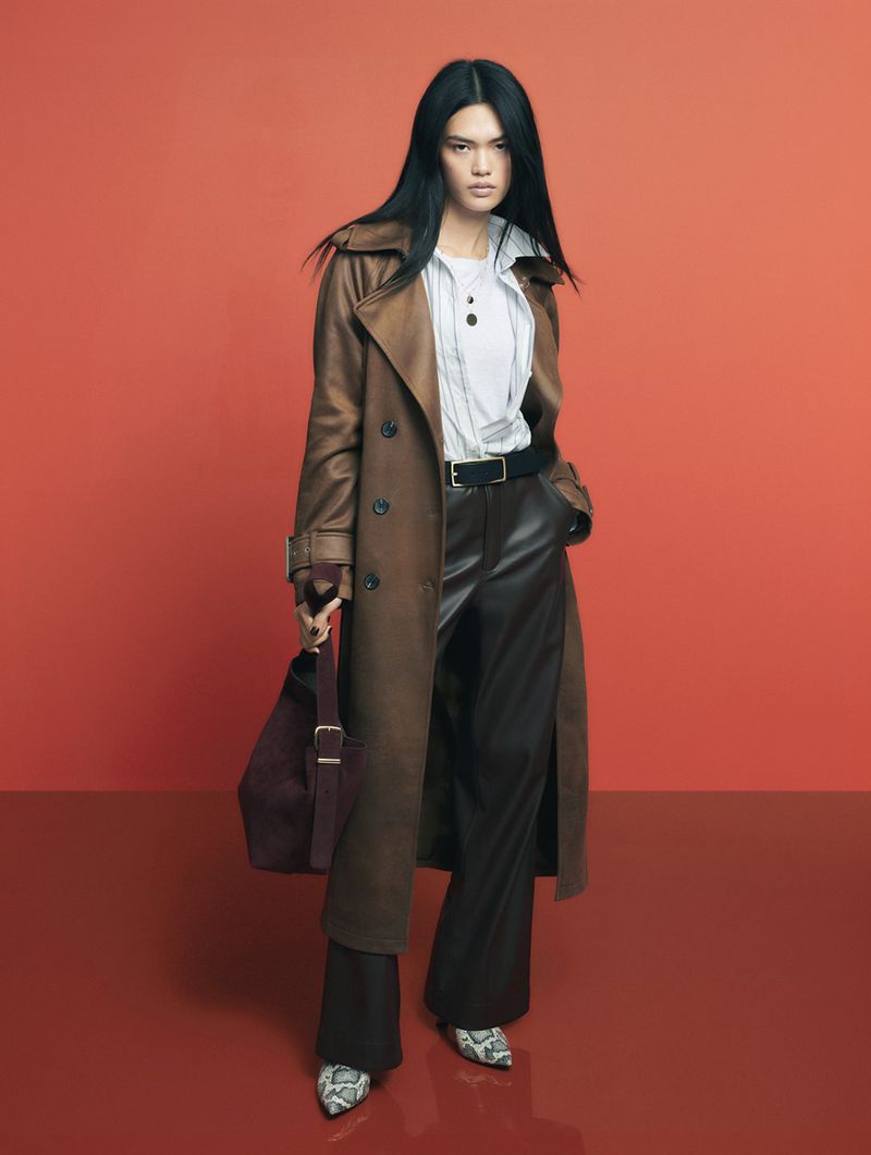 Full-length shot of a woman in a tan trench coat, black pants, and snakeskin shoes, holding a purple bag against a red background.