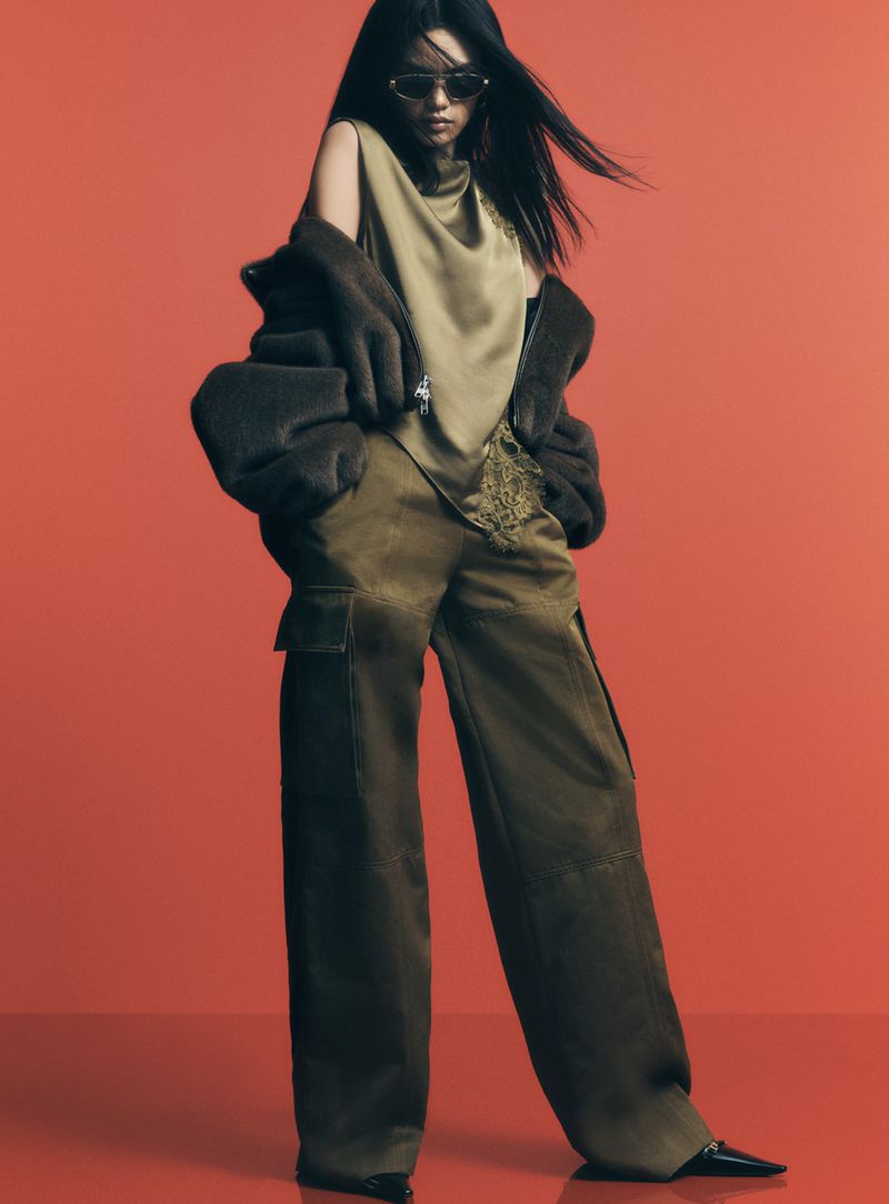 A woman in sunglasses poses in olive green cargo pants, a matching top, and a bomber jacket against a red background in studio lighting.
