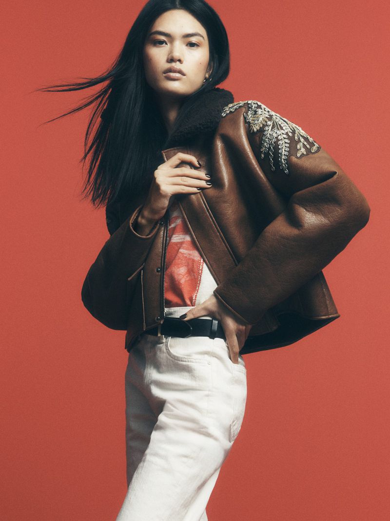 A fashionable Asian woman poses in a brown embroidered shearling jacket, white jeans, and a graphic tee against a red backdrop.
