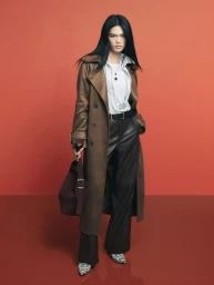 Model Qun Ye wearing a long brown leather coat, dark trousers, and snakeskin boots against a vibrant red backdrop.