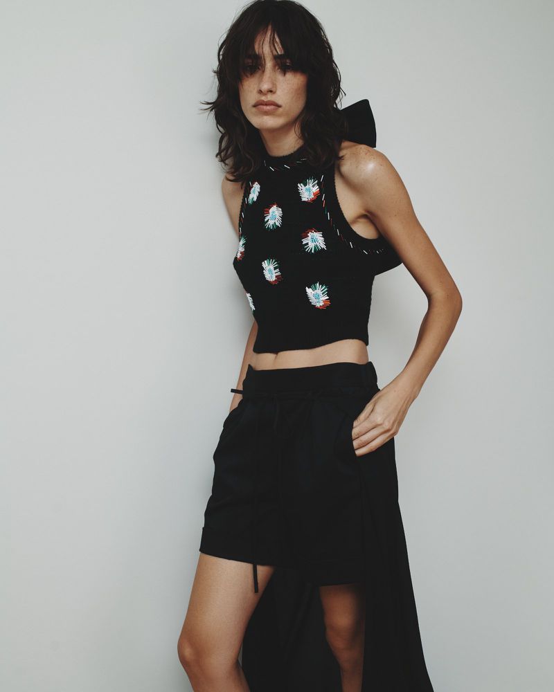 A woman poses in a black crop top with floral embellishments and high-waisted shorts against a plain backdrop, fashion editorial.