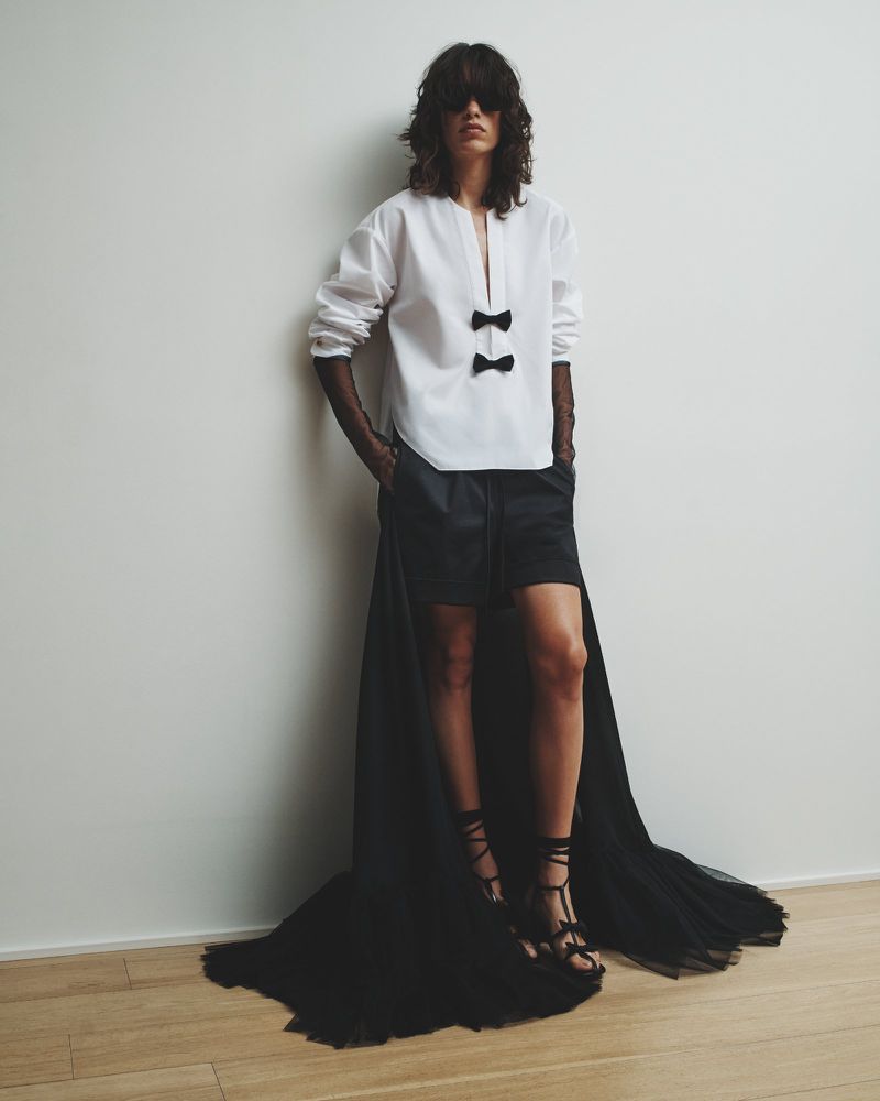 Fashion model poses in a white blouse with bow details, black shorts, and strappy sandals against a neutral background, fashion editorial shot.