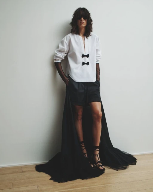 Fashion model poses in a white blouse with bow details, black shorts, and strappy sandals against a neutral background, fashion editorial shot.