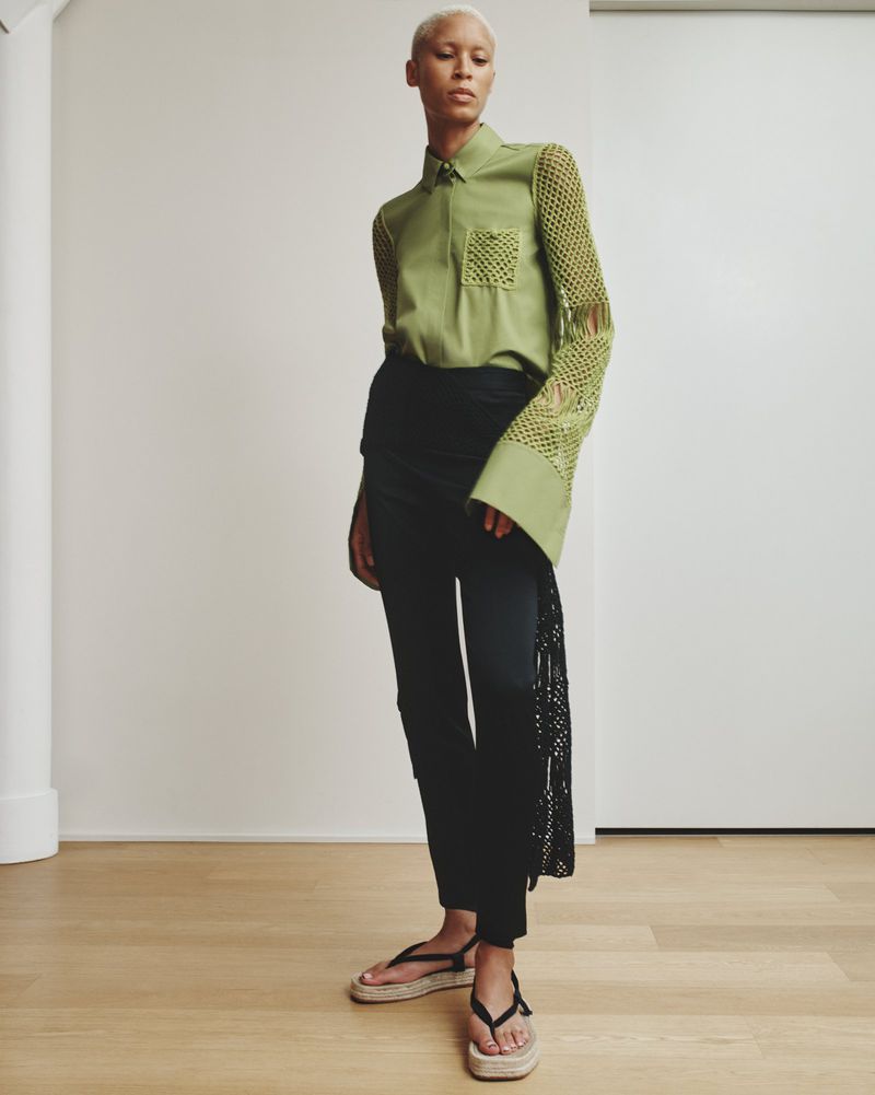 An adult fashion model poses indoors wearing a green collared shirt with crochet sleeves, black pants, and strappy sandals. 