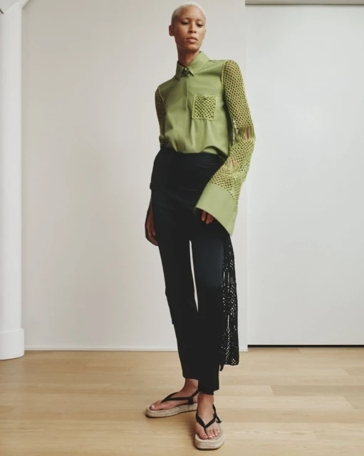 An adult fashion model poses indoors wearing a green collared shirt with crochet sleeves, black pants, and strappy sandals. 