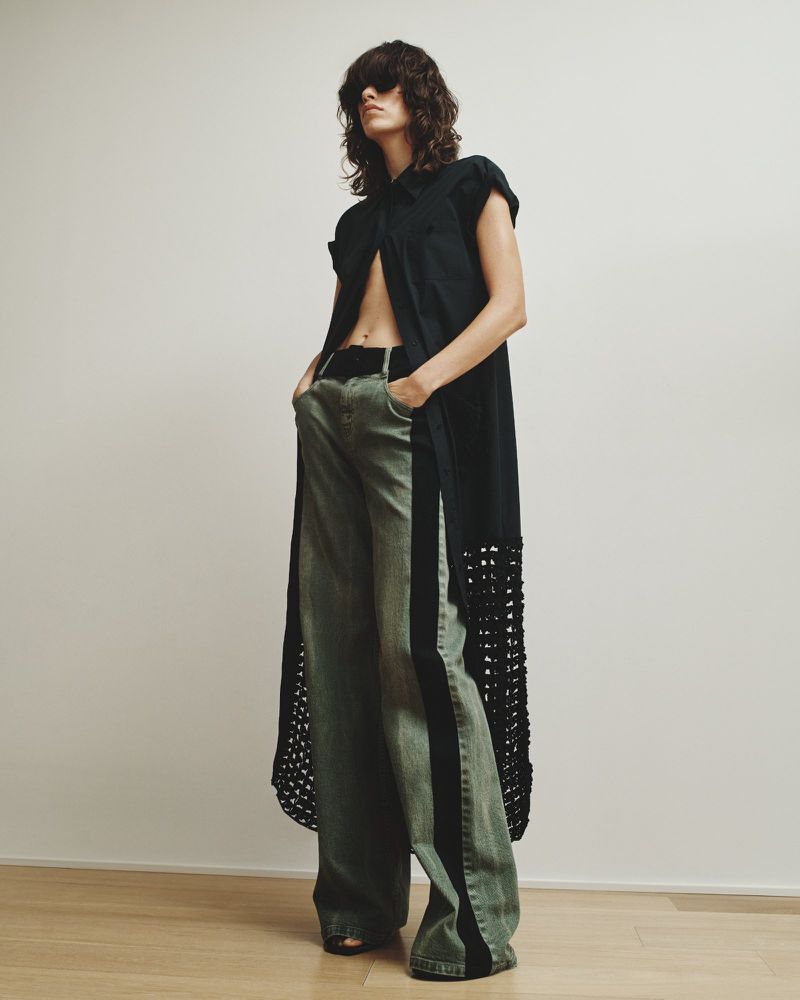 Woman posing indoors in a black sleeveless dress shirt and wide-leg green jeans with black stripe, in a fashion photography style.