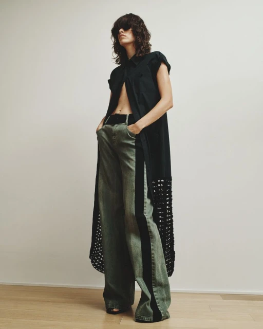 Woman posing indoors in a black sleeveless dress shirt and wide-leg green jeans with black stripe, in a fashion photography style.