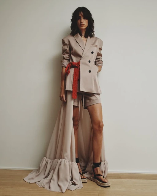 Woman poses in a beige double-breasted blazer, shorts, and sandals in a studio with a reddish-orange belt accenting the look.