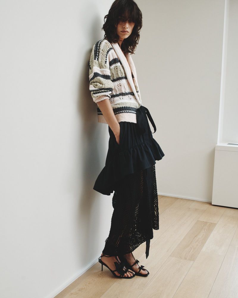 A woman in a knitted cardigan and black skirt leans against a white wall in a full shot, showcasing a minimalist indoor fashion setting.