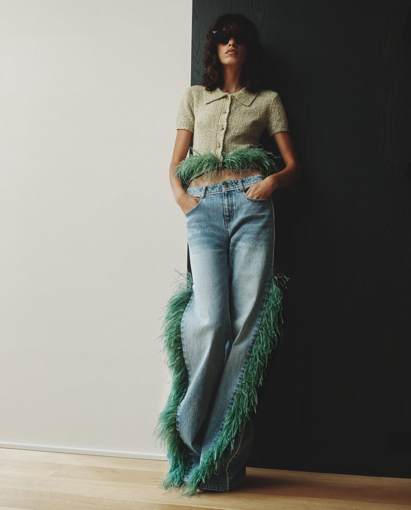 Woman in light-wash, wide-leg jeans with green feather trim and a light green, button-down sweater posing against a wall.