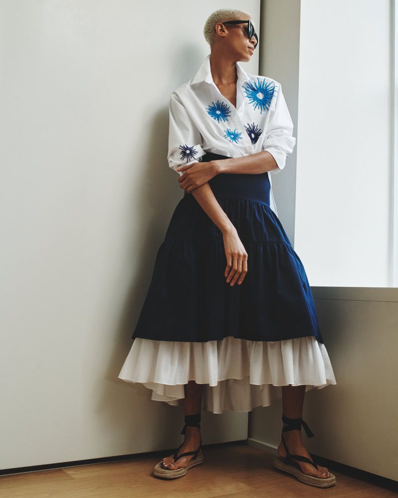 A woman with short hair and sunglasses models a white collared shirt with blue floral embroidery and a navy and white tiered skirt.