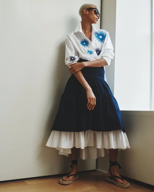 A woman with short hair and sunglasses models a white collared shirt with blue floral embroidery and a navy and white tiered skirt.