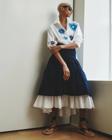 A woman with short hair and sunglasses models a white collared shirt with blue floral embroidery and a navy and white tiered skirt.