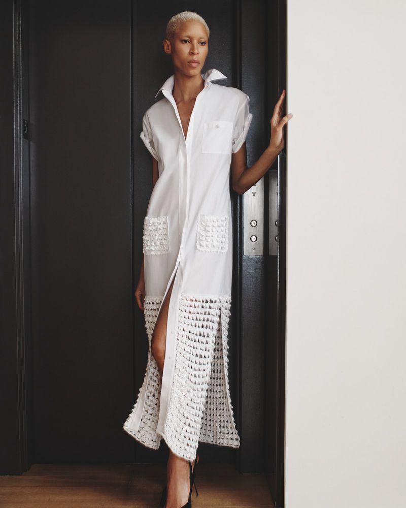 Elegant adult female poses in a white dress with unique crochet details, standing in front of a grey elevator in an indoor setting.