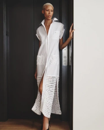 Elegant adult female poses in a white dress with unique crochet details, standing in front of a grey elevator in an indoor setting.