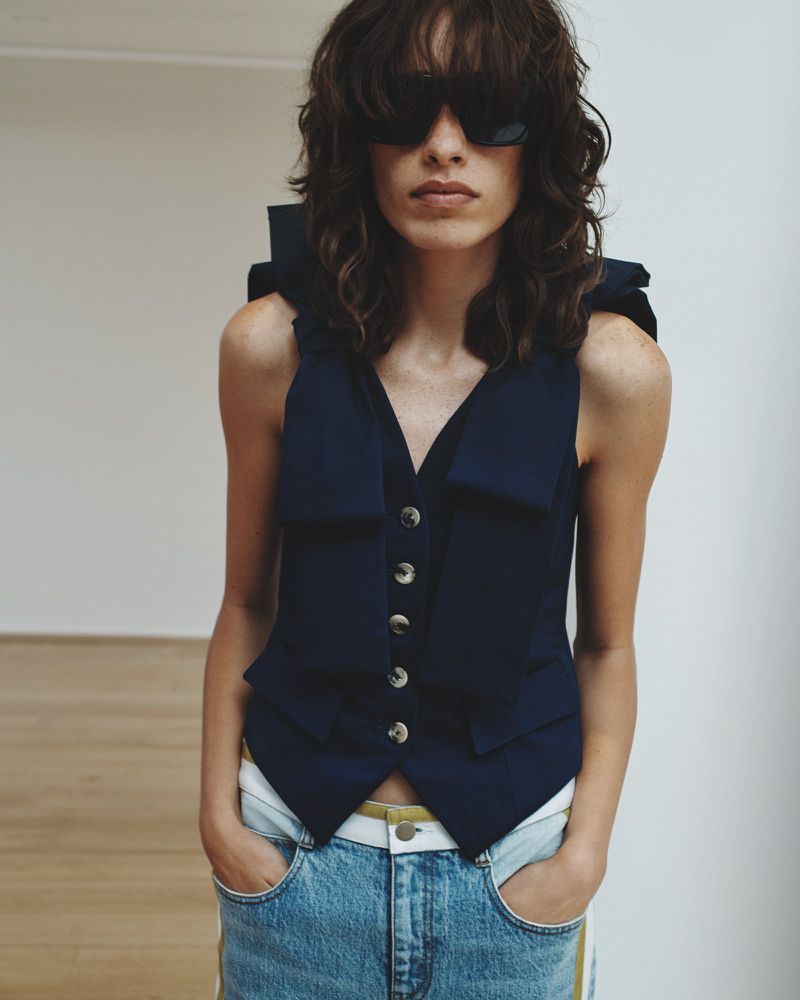 A woman wearing a sleeveless navy vest and light-wash jeans with her hands in her pockets poses in a minimalist studio.