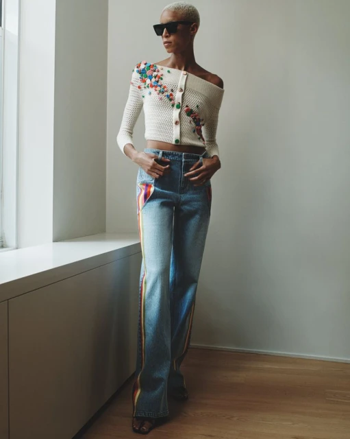 A woman with short blonde hair poses indoors wearing an off-the-shoulder knit top and jeans with rainbow stripe accents.