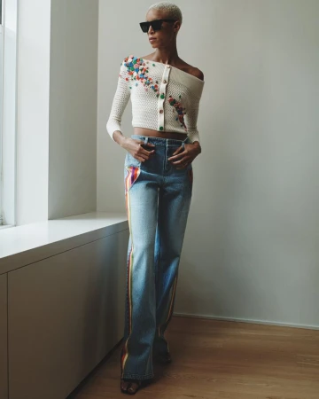 A woman with short blonde hair poses indoors wearing an off-the-shoulder knit top and jeans with rainbow stripe accents.